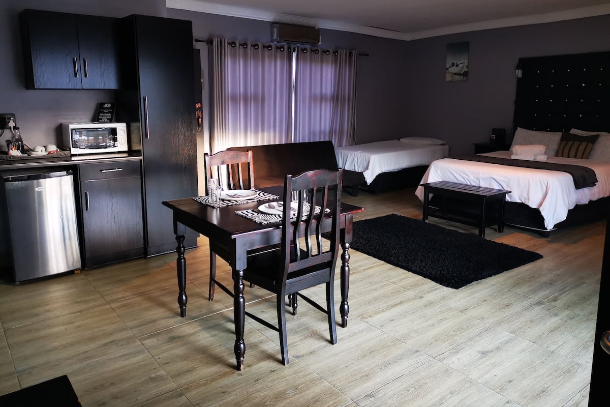 A compact hotel room with a kitchenette, a dining area, two single beds, and a small desk.
