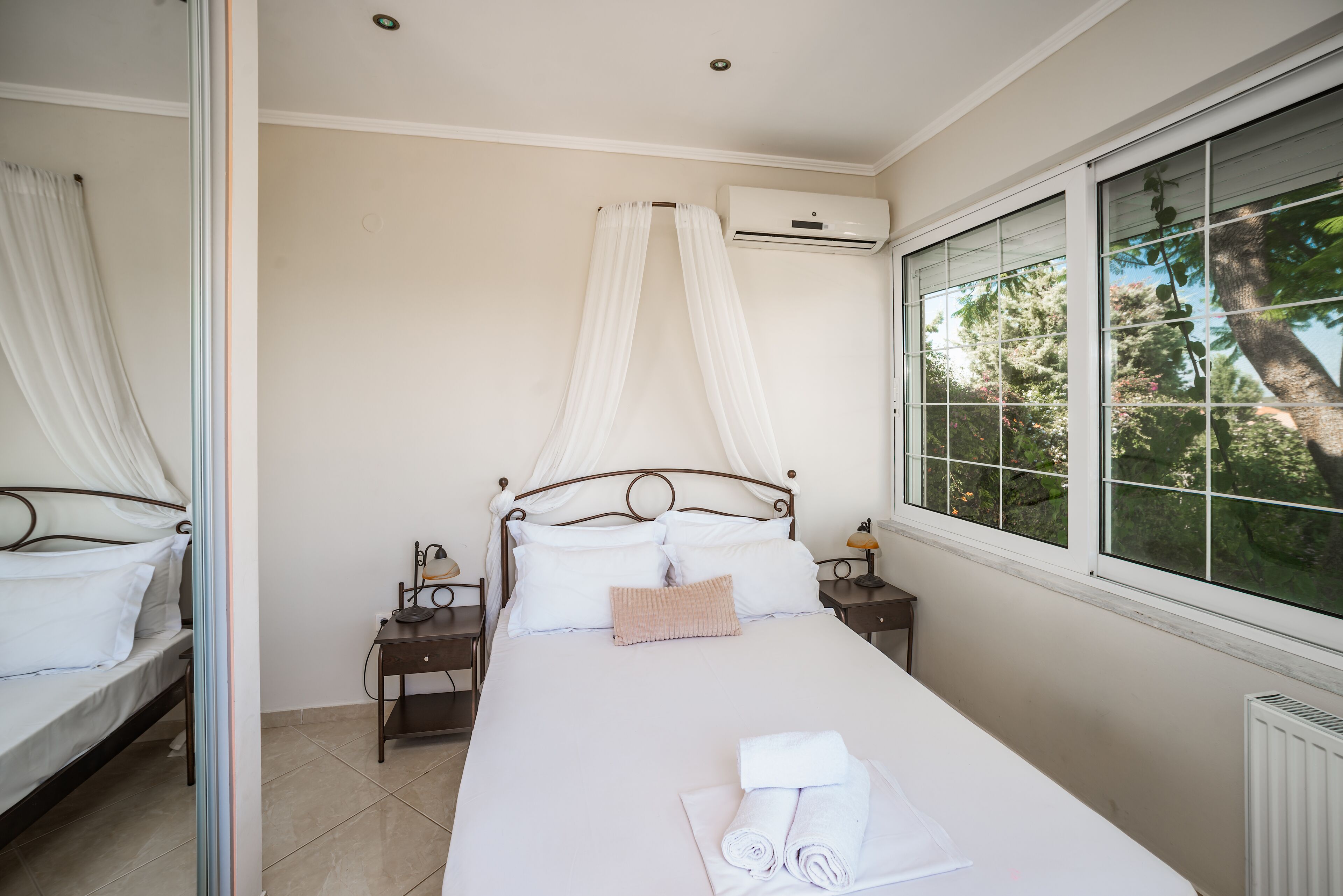 Un dormitorio con cama, mesitas de noche, un aire acondicionado y una ventana con vistas a árboles.