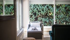 A spa area with a massage table, two woven ottomans, and a view of a dense green forest through large windows.