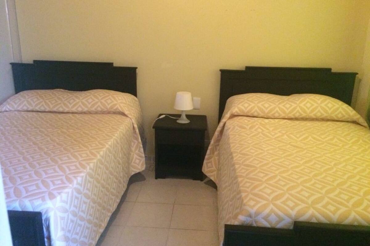 A room with two beds, each with a patterned bedspread and a dark headboard. There is a small bedside table with a lamp between the beds.