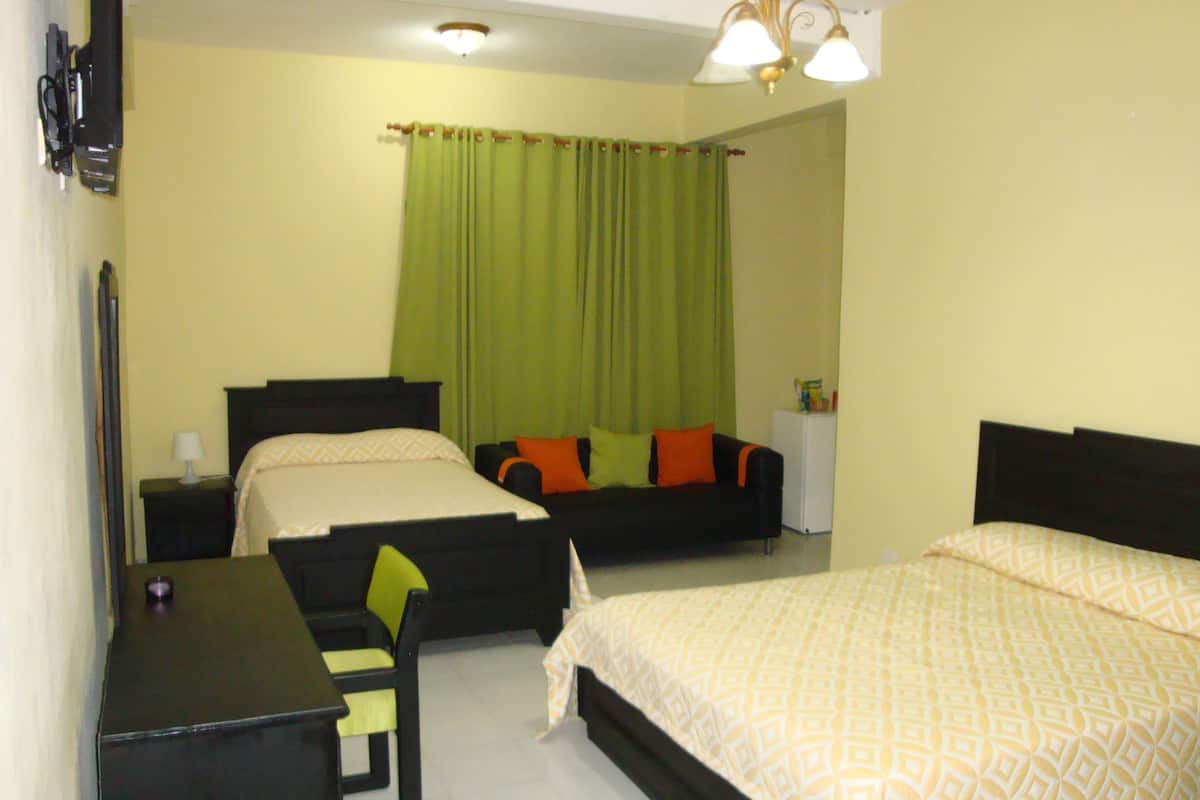 A hotel room with two beds, a desk, a chair, a small table, a nightstand, a lamp, a refrigerator, and a curtain.