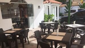Un área de café al aire libre con mesas y sillas, una puerta de vidrio de entrada y un edificio blanco con tejado de tejas rojas al fondo.