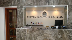 Mostrador de recepción en el Hotel Santander, S.D., con una pared de mármol, un reloj, banderas y un monitor de computadora.