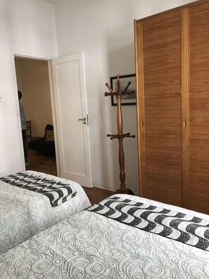 A bedroom with two beds, a wooden wardrobe, a coat rack, and a door leading to another room.