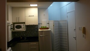 Cocina compacta con armarios blancos, microondas y refrigerador. Encima del lavamanos, hay un estante con objetos decorativos.