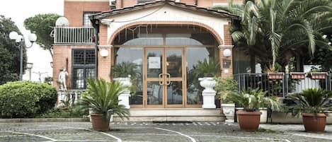 A hotel entrance with a brick facade, a glass door, and a sign indicating it is a hotel. There are potted plants and a cobblestone pathway leading to the entrance.