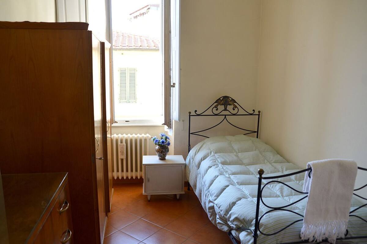 A bedroom with a bed, a radiator, a small bedside table with a vase of flowers, and a wooden wardrobe.
