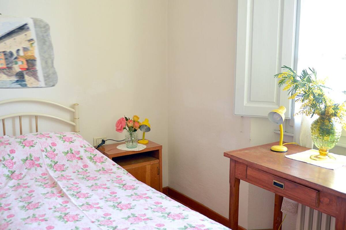 A bedroom with a bed, a bedside table with a lamp, a small desk, and a window with a view.