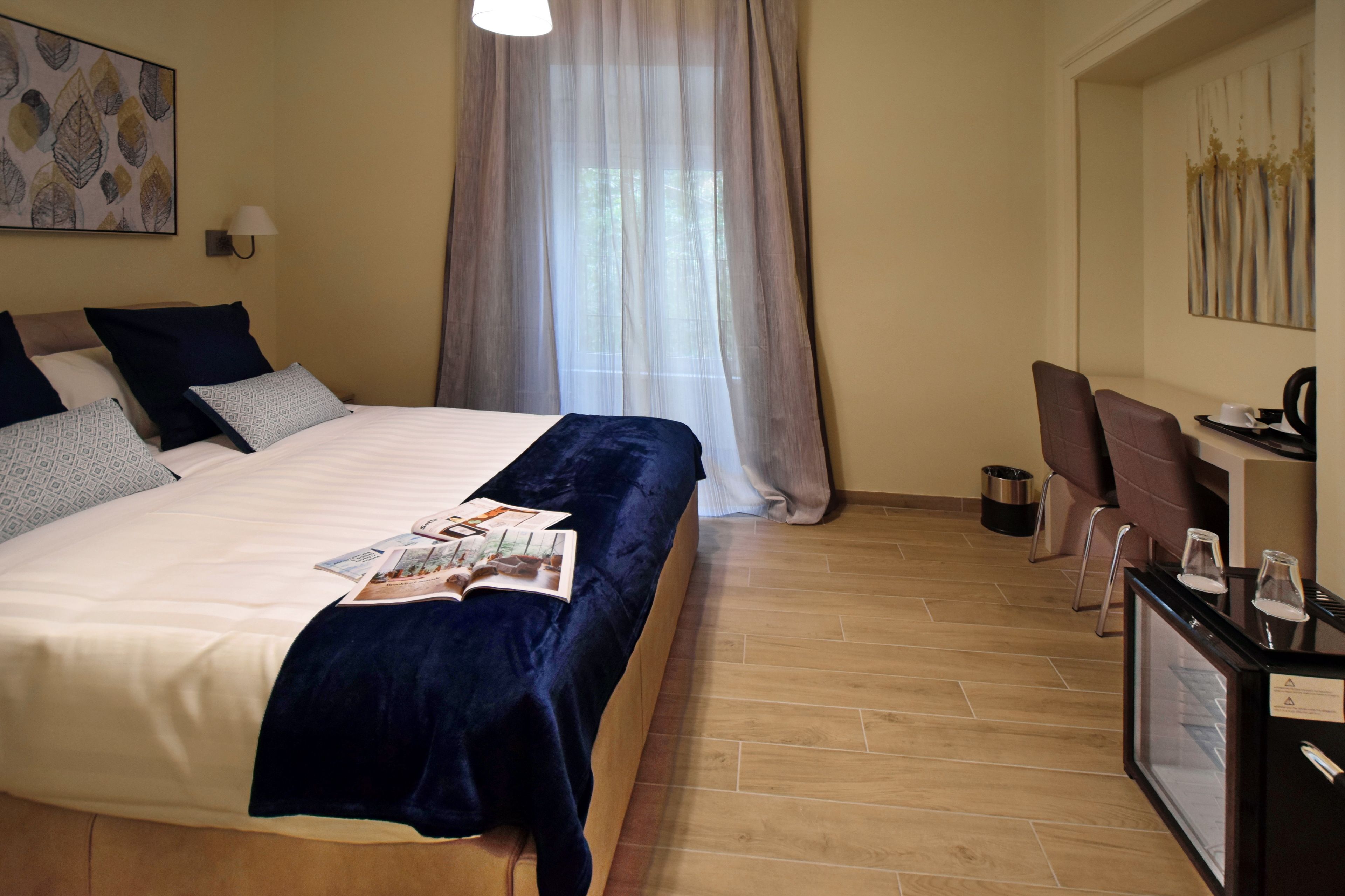 Quarto de hotel com uma cama grande, uma escrivaninha com cadeira, uma mesinha com cafeteira e uma janela com cortinas leves.