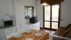 Un coin repas avec une table dressée pour quatre personnes, un four à micro-ondes, un téléviseur posé sur un meuble et une porte-fenêtre coulissante donnant sur un balcon.