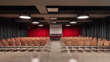 A theater with rows of chairs, a stage with a red curtain, and overhead lighting.