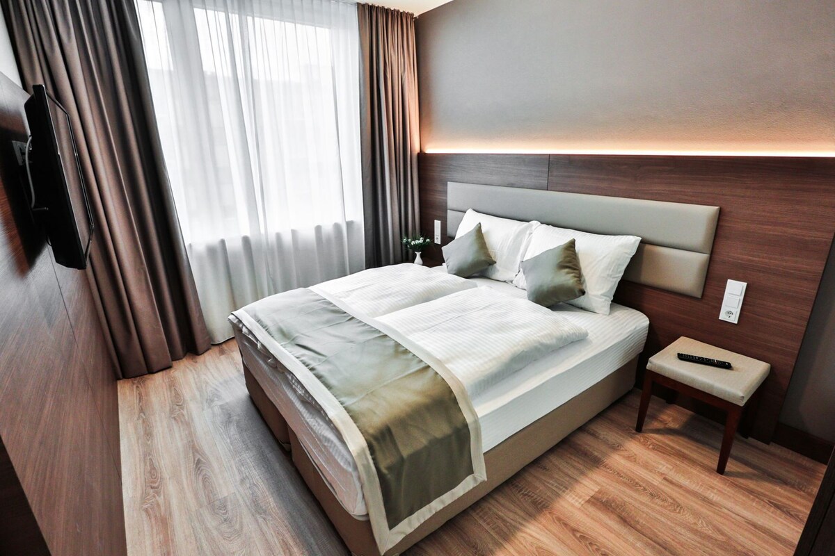 Una habitación de hotel moderna con una cama grande, una mesita de noche, un televisor montado en la pared y una ventana con cortinas.