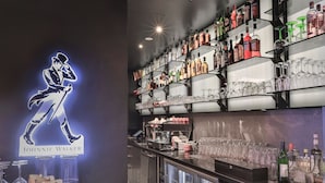 Een bar met planken vol drankflessen en glazen, een koffieapparaat, en een neonbord met een dansende figuur.