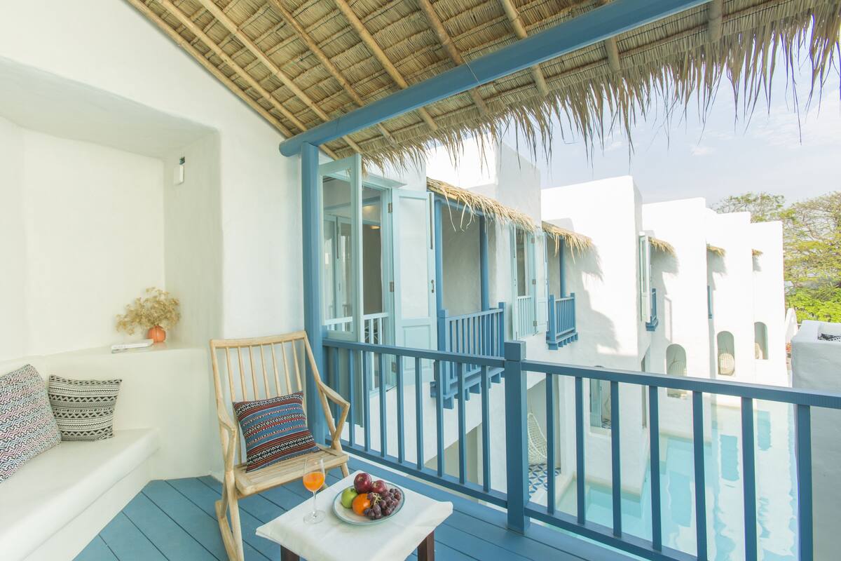 Un balcon doté d’un toit de chaume, une chaise en bois, une table avec des fruits, et une vue sur une piscine et des bâtiments blancs.