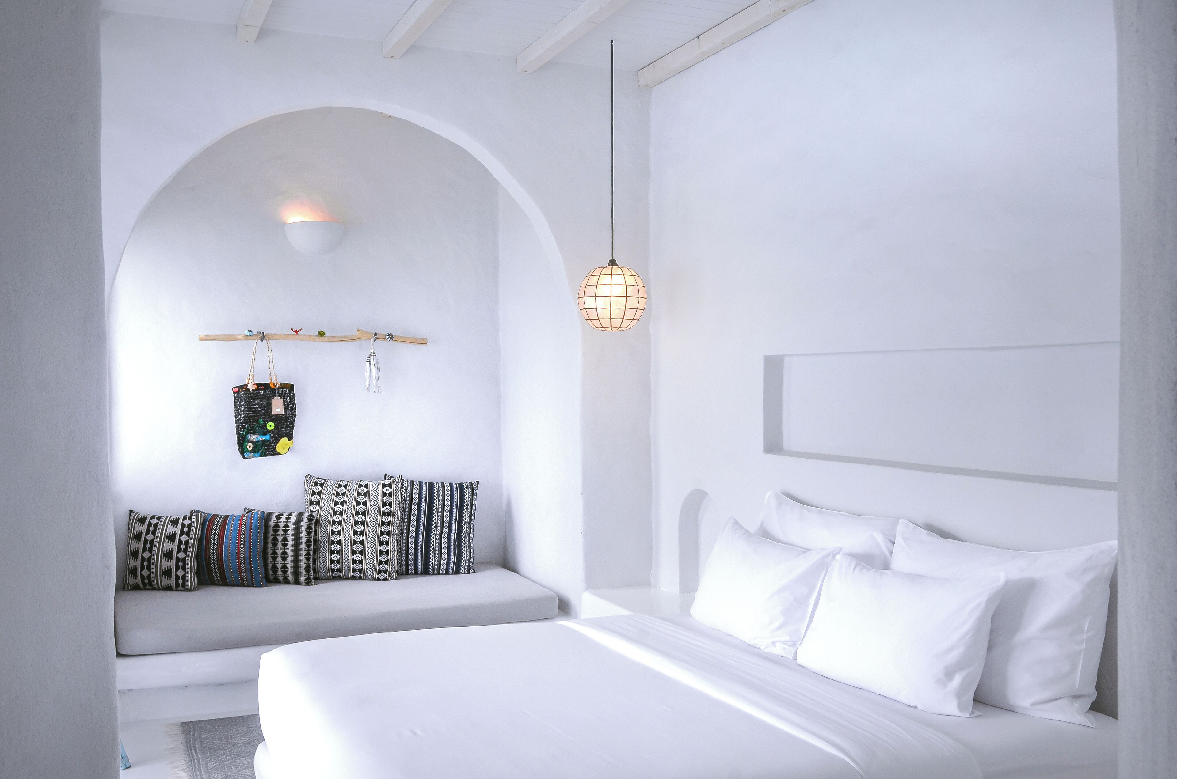 A minimalist white room with a bed, a bench, and a hanging light fixture.