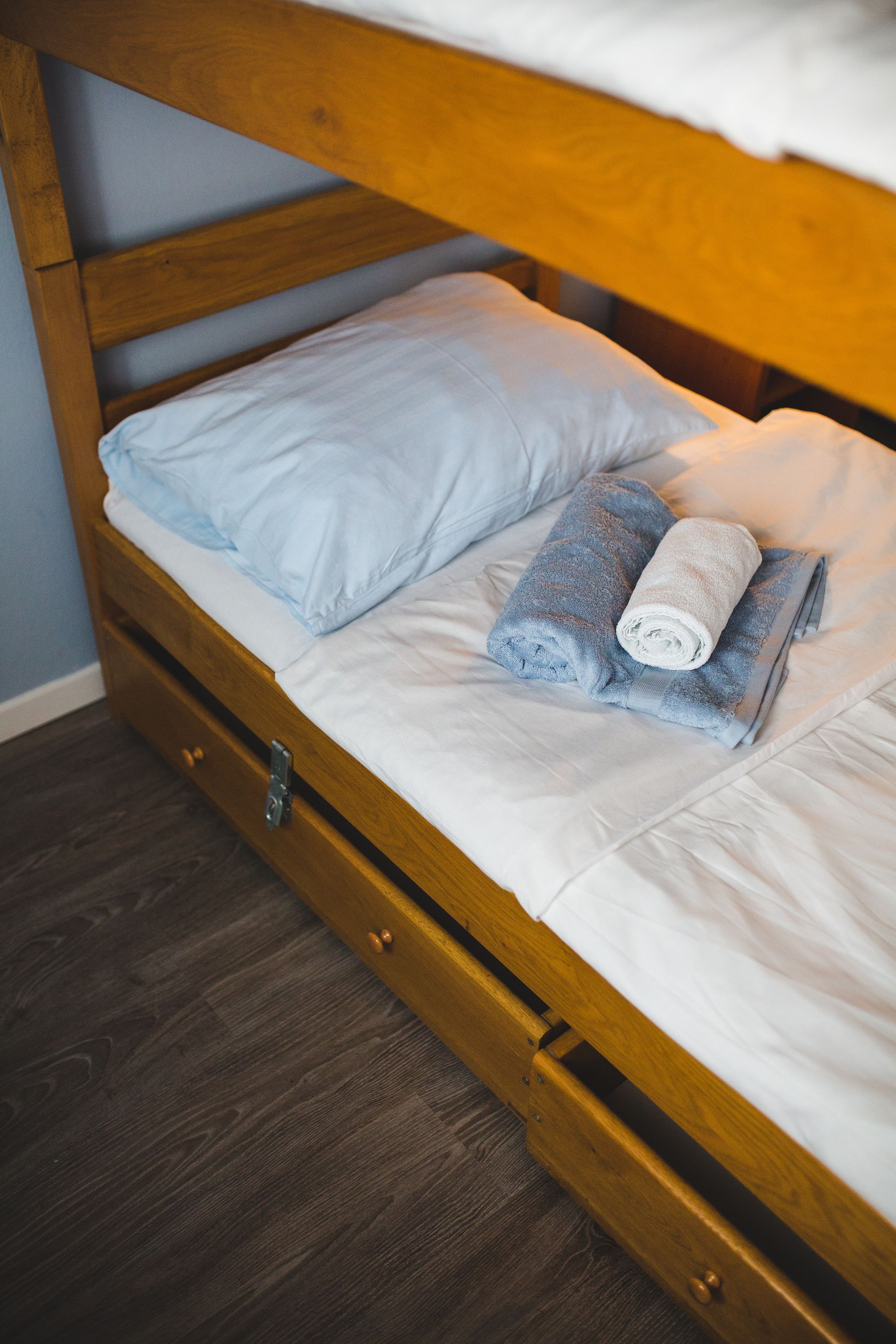 A wooden bunk bed with a white mattress and a single blue pillow. There are two rolled towels on the bed.