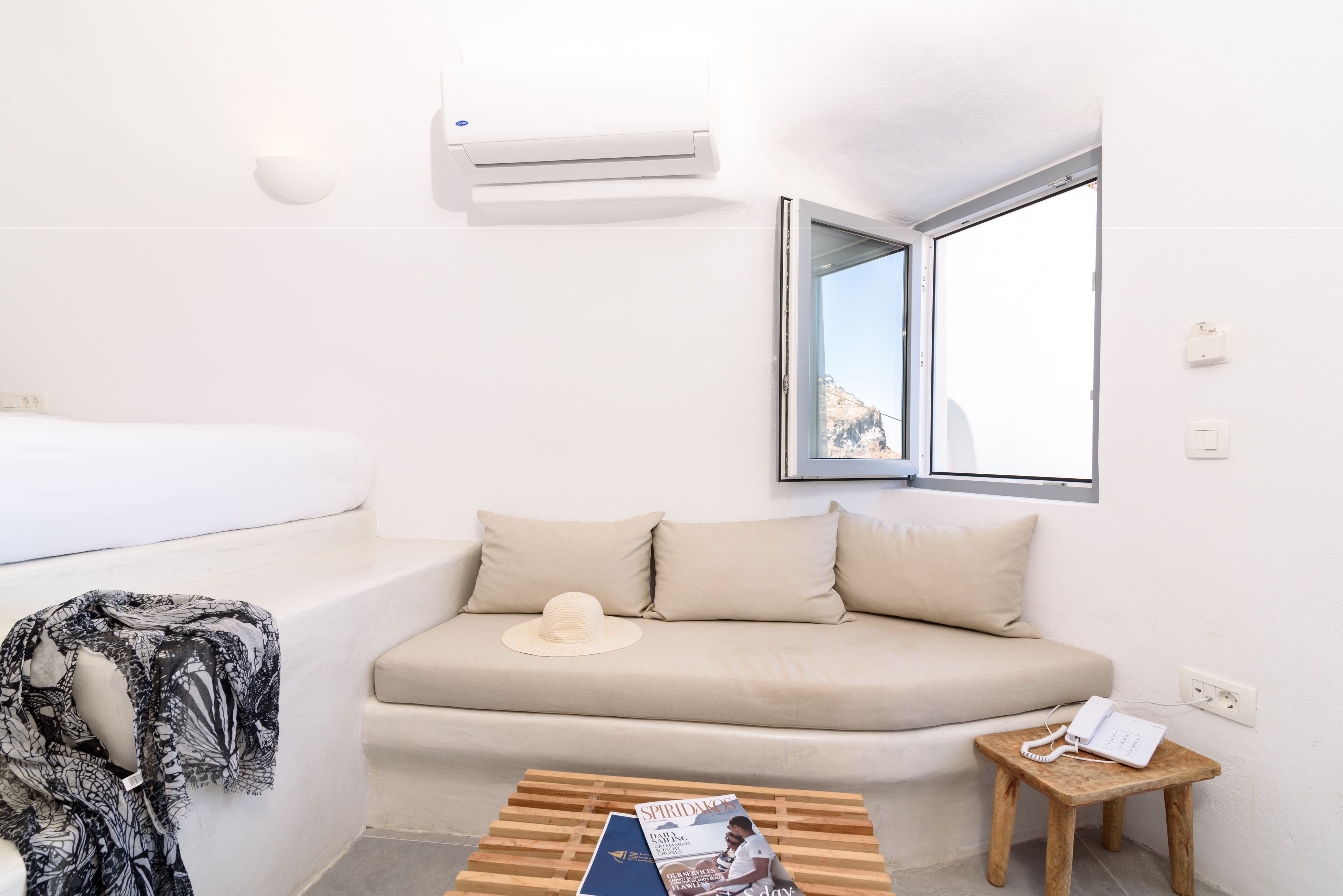 A modern, minimalist hotel room with a sofa bed, a small wooden table with a magazine, a telephone, and a window with a mountain view.