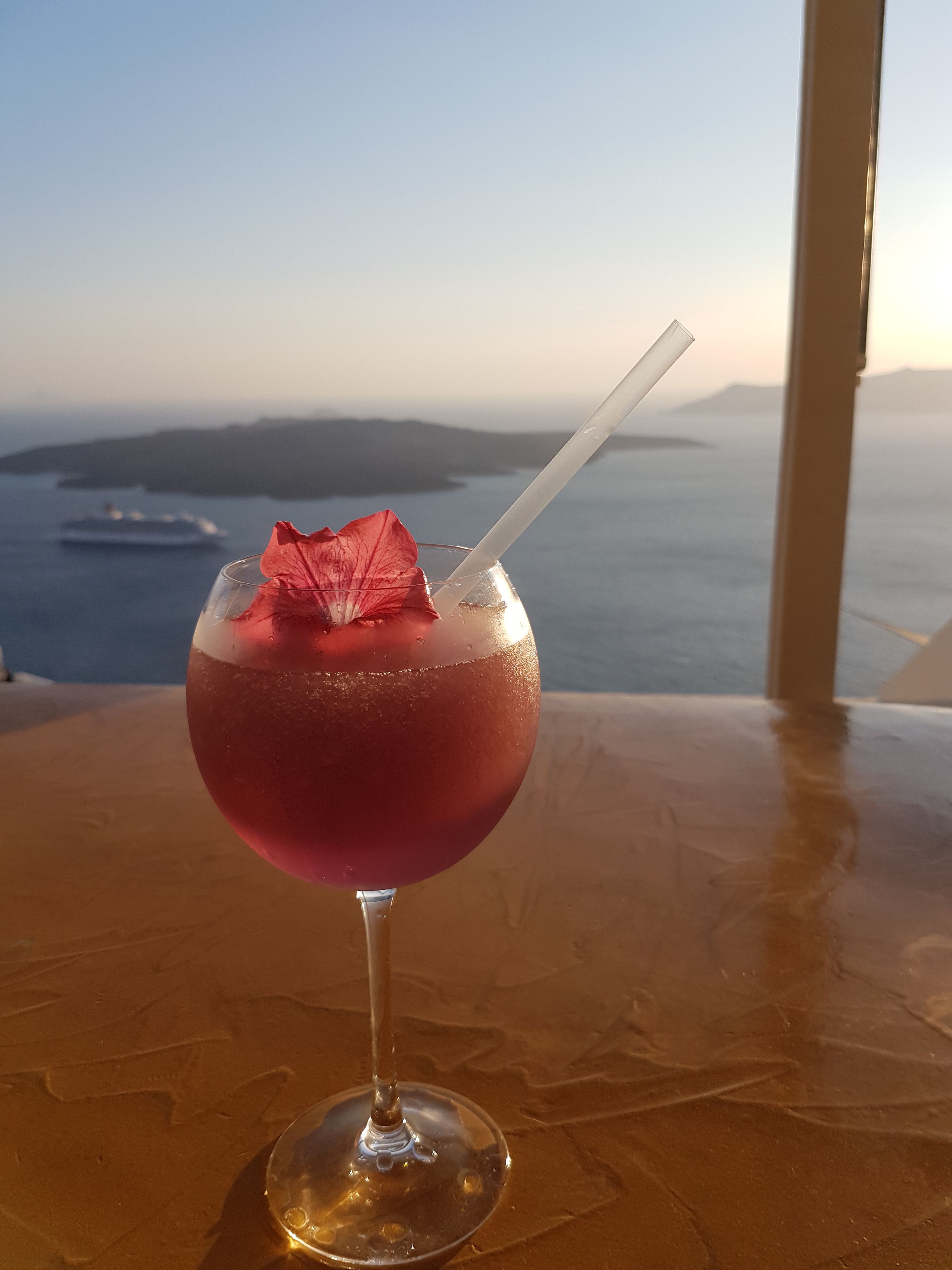 A cocktail with a straw and a decorative leaf on a table with a view of the sea and a ship in the distance.