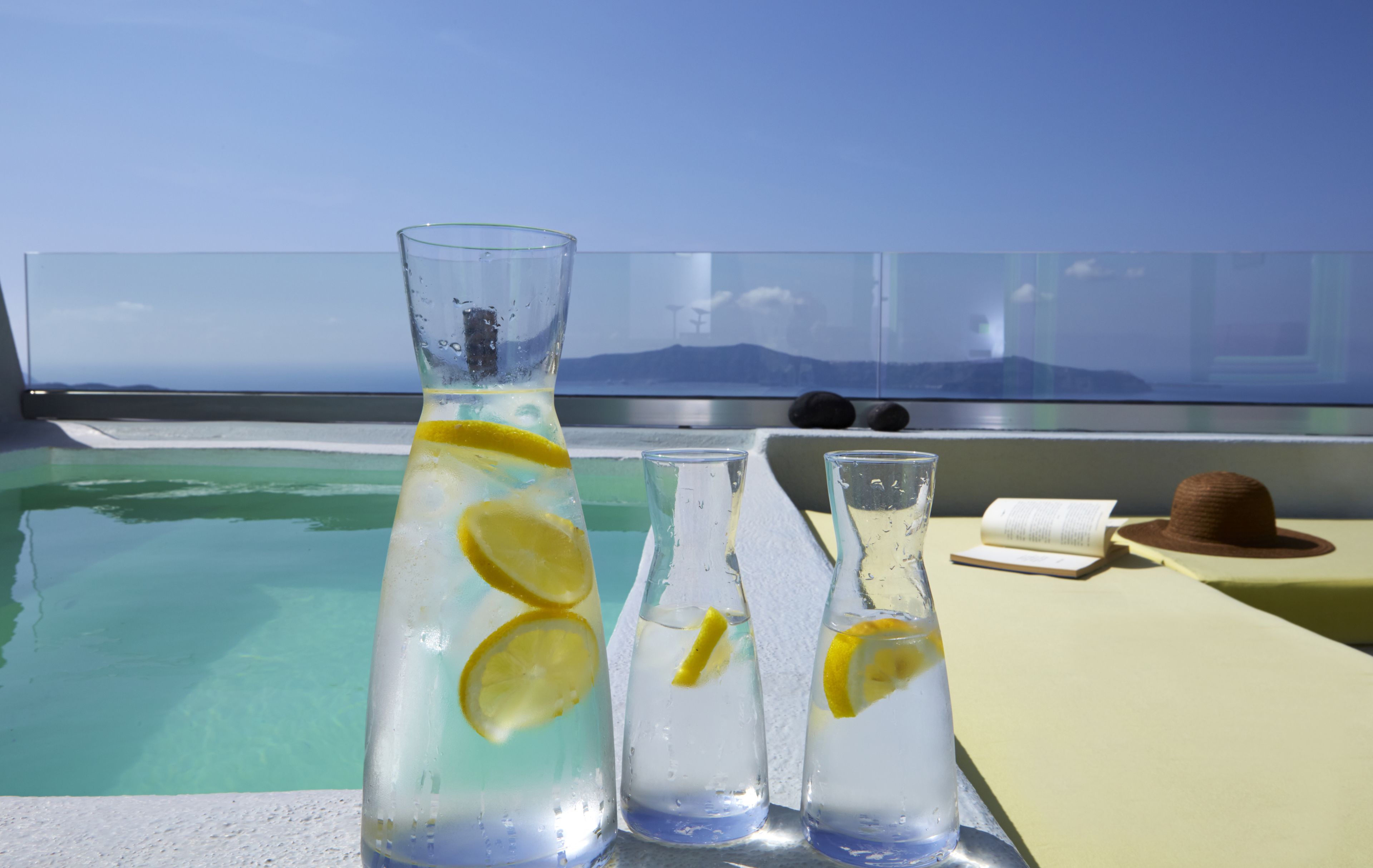 Ein klarer Glasbehälter und zwei kleinere Gläser mit Zitronenscheiben auf einem Tisch in Poolnähe, mit Blick auf ferne Berge.