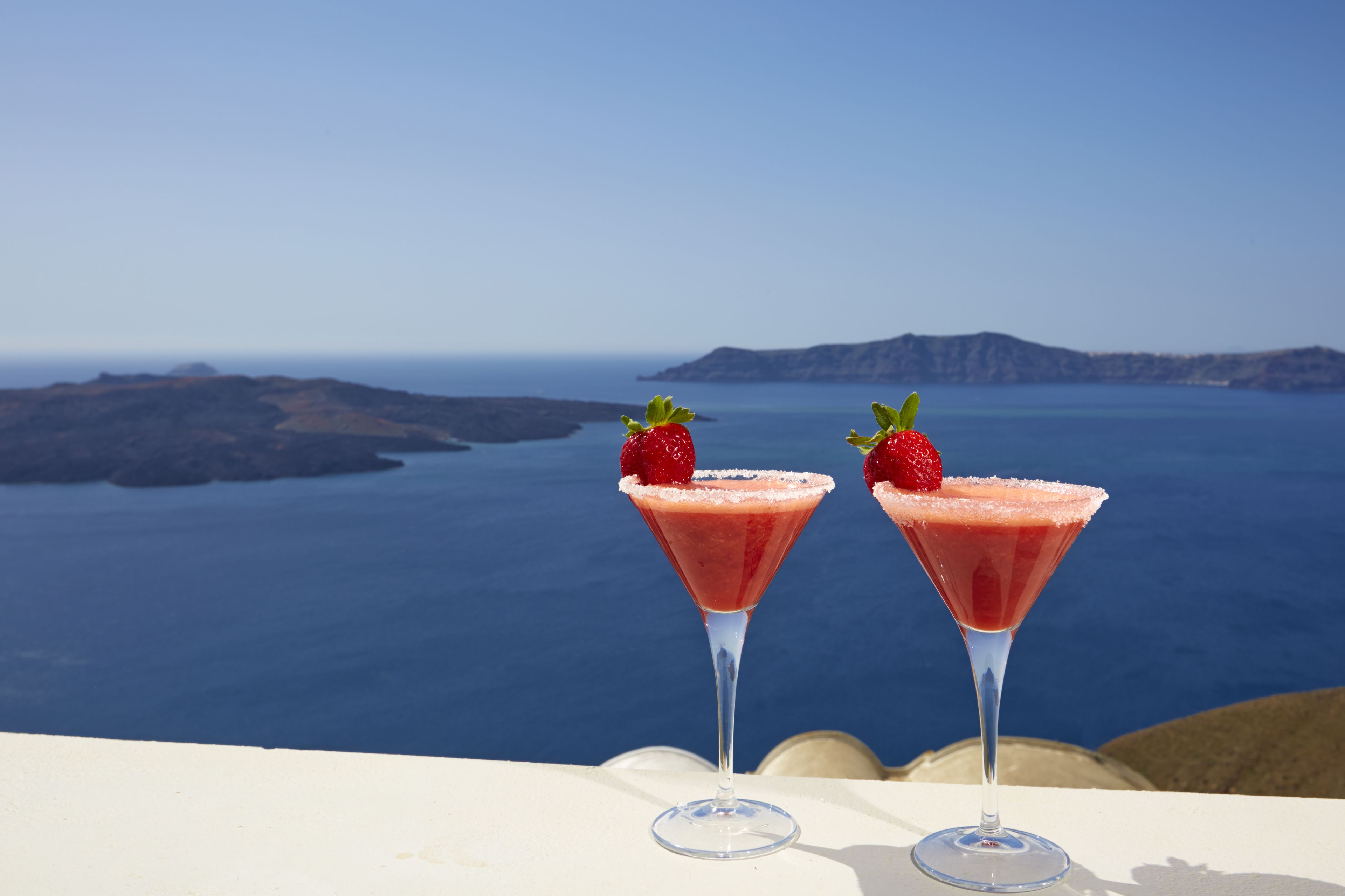 Dos copas de martini con una bebida roja y fresas sobre una mesa con vistas al mar y a las islas.