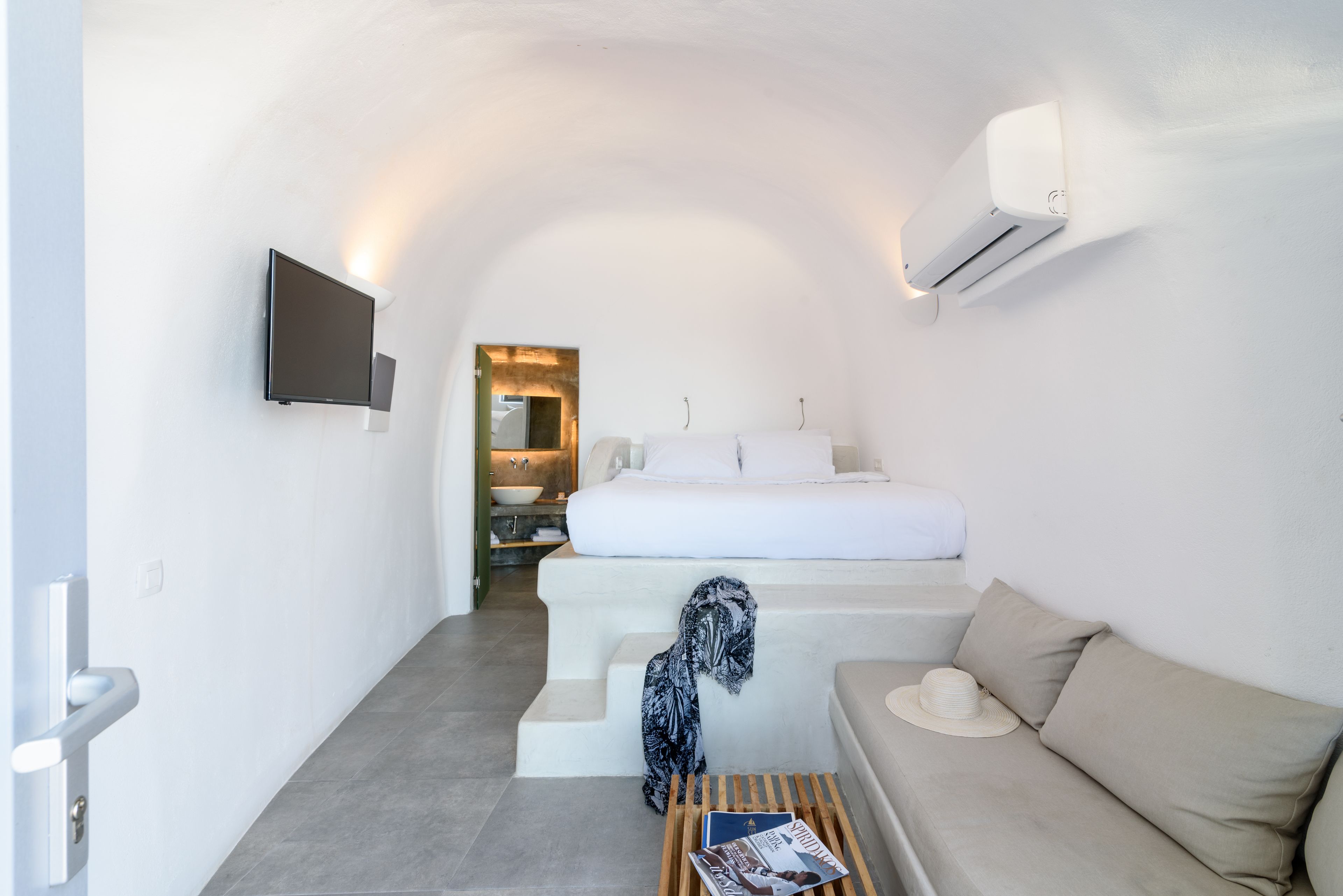 A compact, modern room with a bed, a sofa, a small table with a magazine, and a TV mounted on the wall.