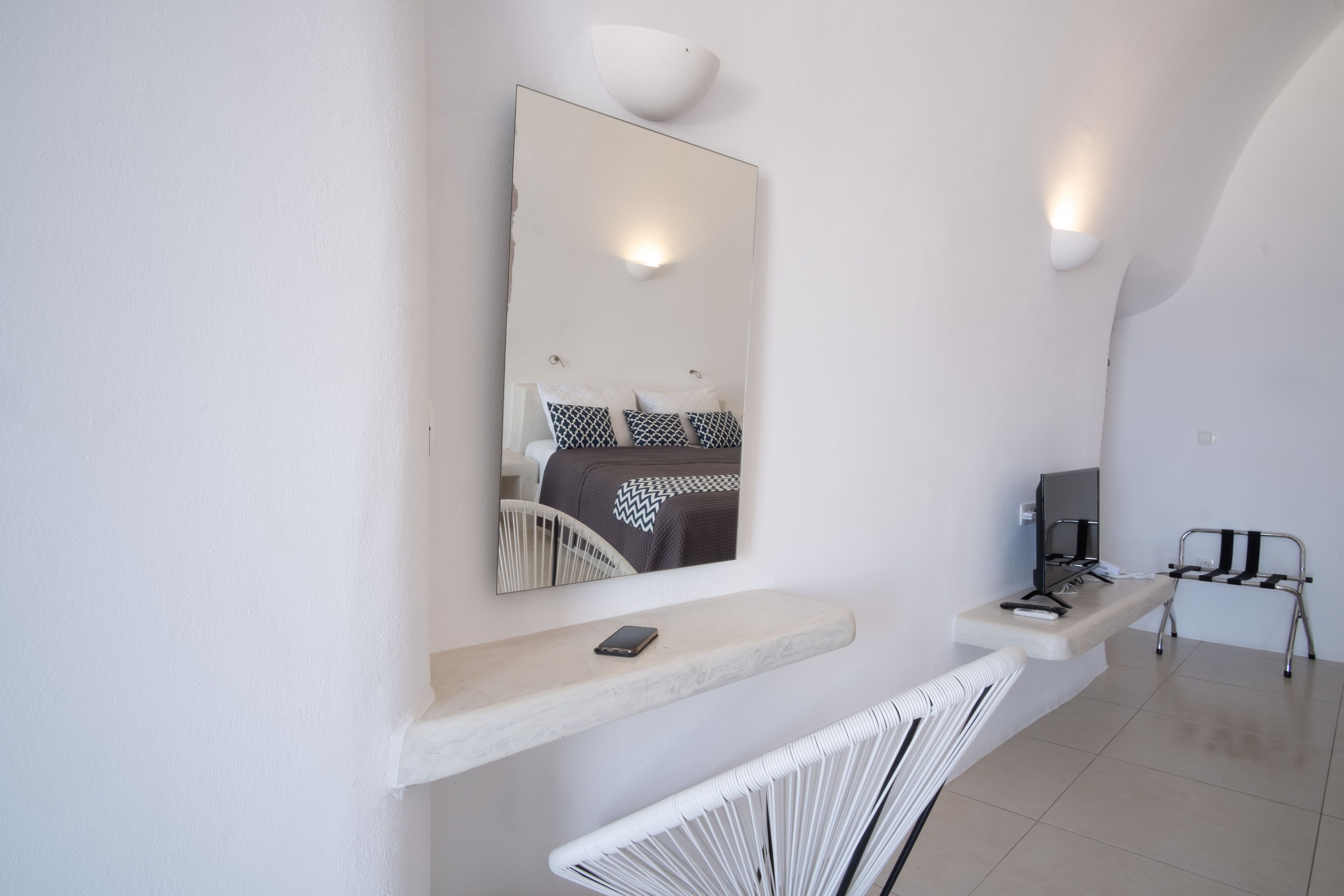 Une chambre à coucher moderne et minimaliste, dotée d’un grand miroir, d’une table de chevet avec un téléphone, d’une chaise et d’un téléviseur.
