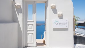 A white door leading to a balcony with a view of the sea. A sign on the wall reads "On The Cliff Suites." - On the Cliff (Santorini)