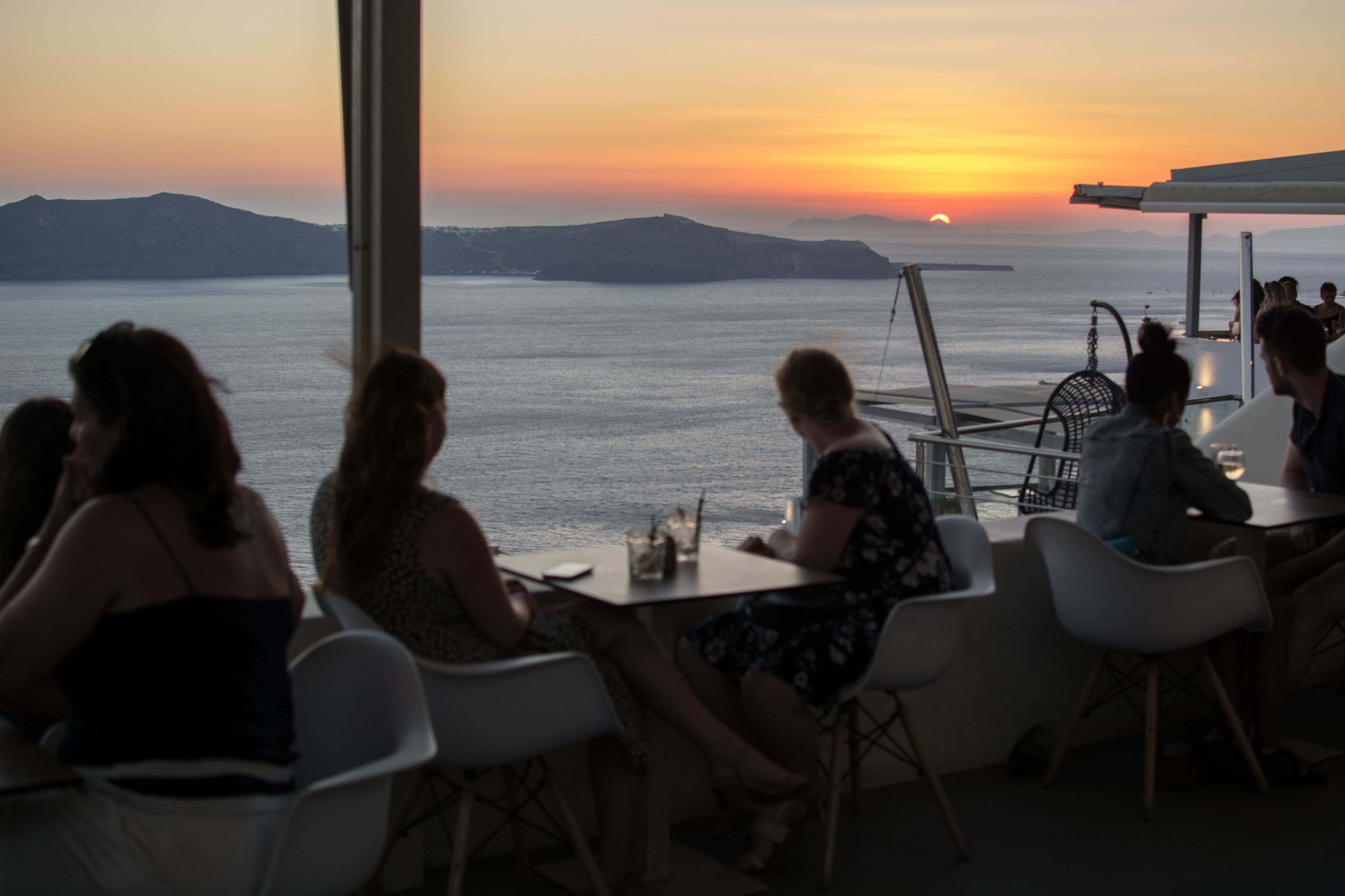 A group of people seated at tables by a window overlooking a body of water during sunset.