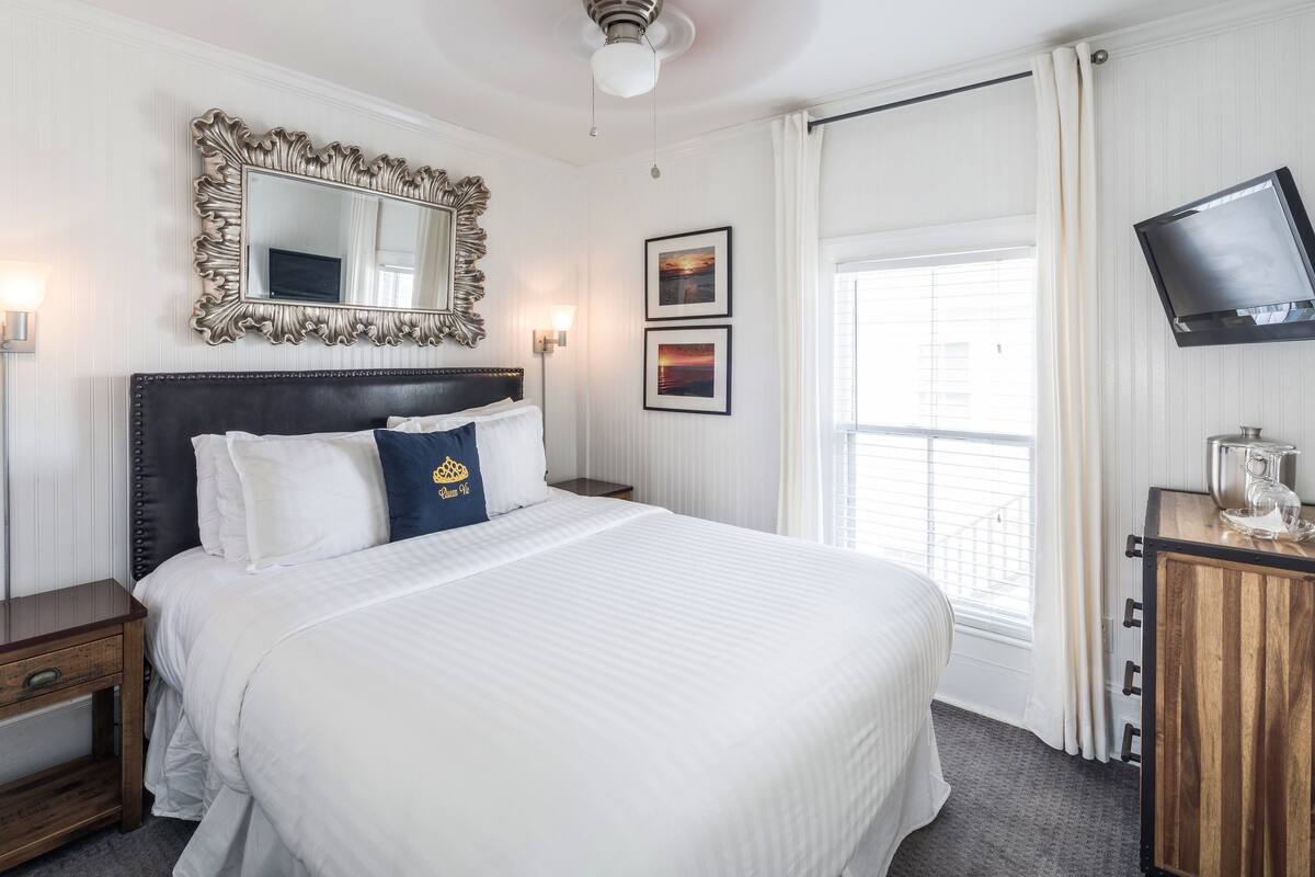 A neatly arranged hotel room with a large bed, a mirror, a flat-screen TV, and a window with curtains.