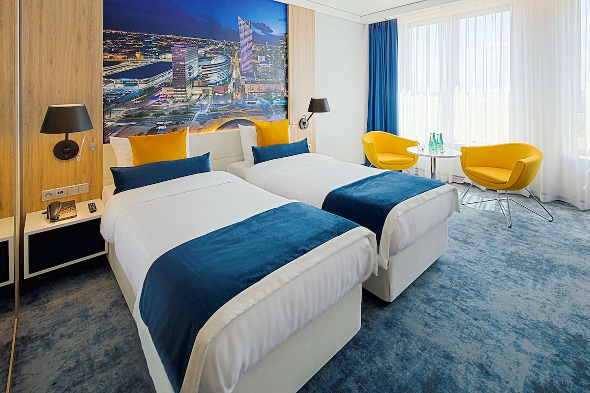 Hotelkamer met twee bedden, een stadsmuur, een kleine tafel met stoelen en uitzicht op de stad door grote ramen.
