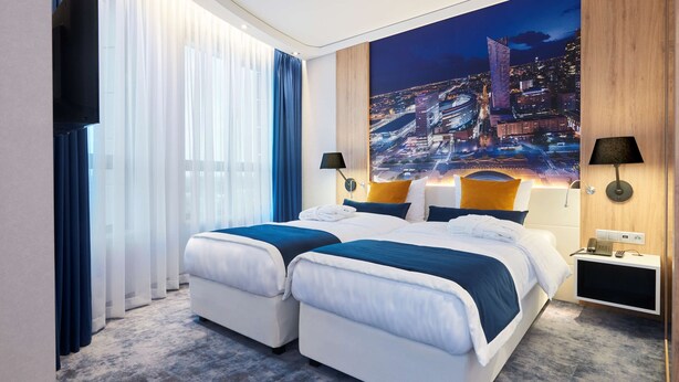 Habitación de hotel con dos camas, un mural grande de un paisaje urbano, un televisor de pantalla plana y una mesita de noche con lámpara.