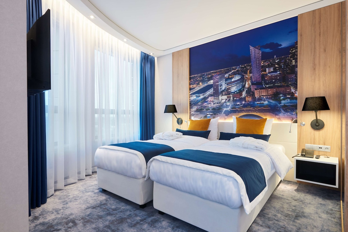 Une chambre d’hôtel avec deux lits, une grande fresque représentant un paysage urbain, un téléviseur à écran plat et une table de chevet avec une lampe.