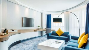 Una sala de estar moderna con distribución curva, un televisor de pantalla plana en la pared, un sofá azul con cojines amarillos, una mesa de centro blanca y una lámpara de pie.