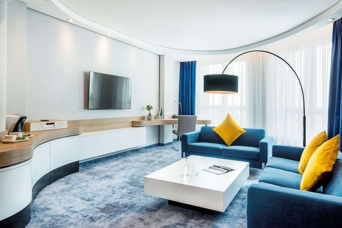 Een moderne woonkamer met een gebogen indeling, een flatscreen televisie aan de muur, een blauwe bank met gele kussens, een witte salontafel en een vloerlamp.