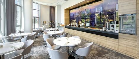 A modern hotel lobby with round tables and chairs, a cityscape mural, and a bar area with kitchen appliances.