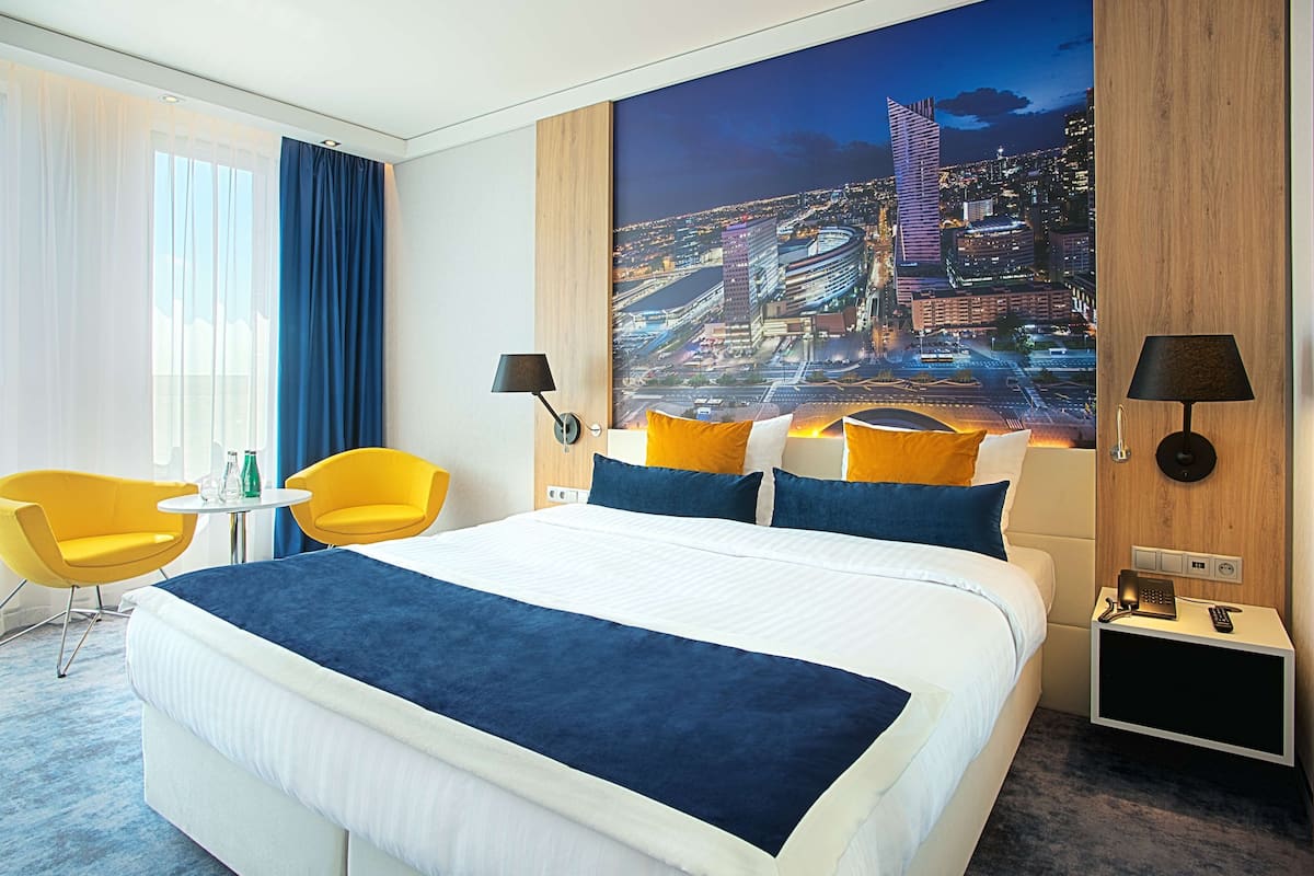Hotelkamer met een groot bed, twee gele stoelen, een klein tafeltje met drankjes, een nachtkastje met een lamp, en een stadsgezicht op de muur.