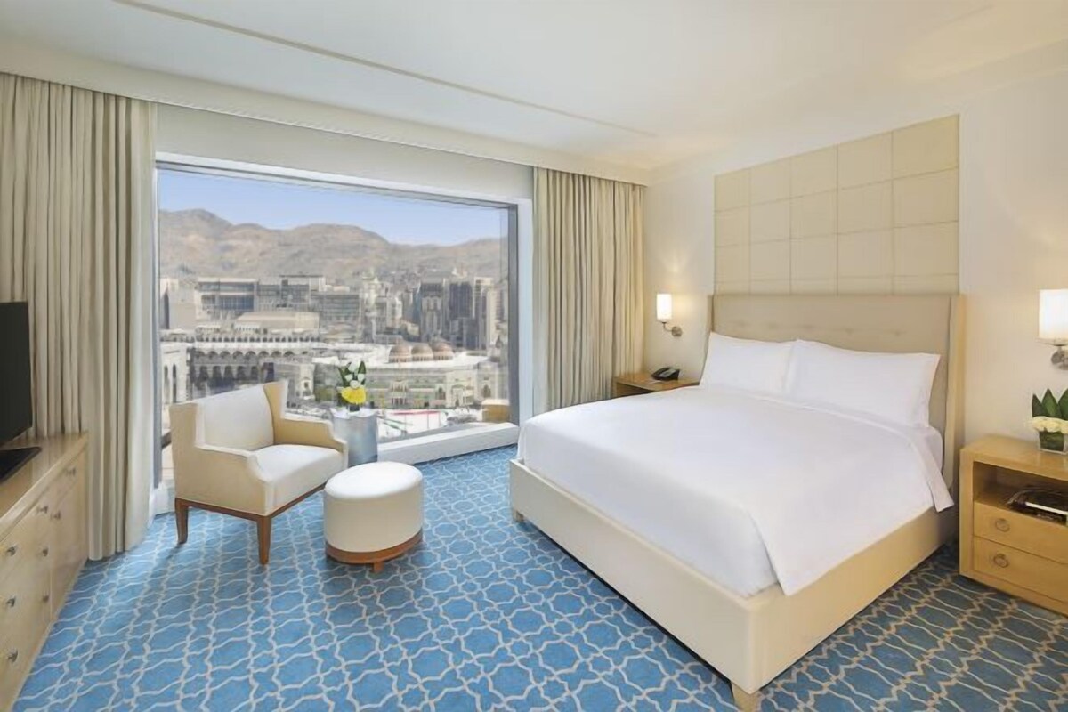 A hotel room with a large bed, a TV, a seating area, and a view of a cityscape.