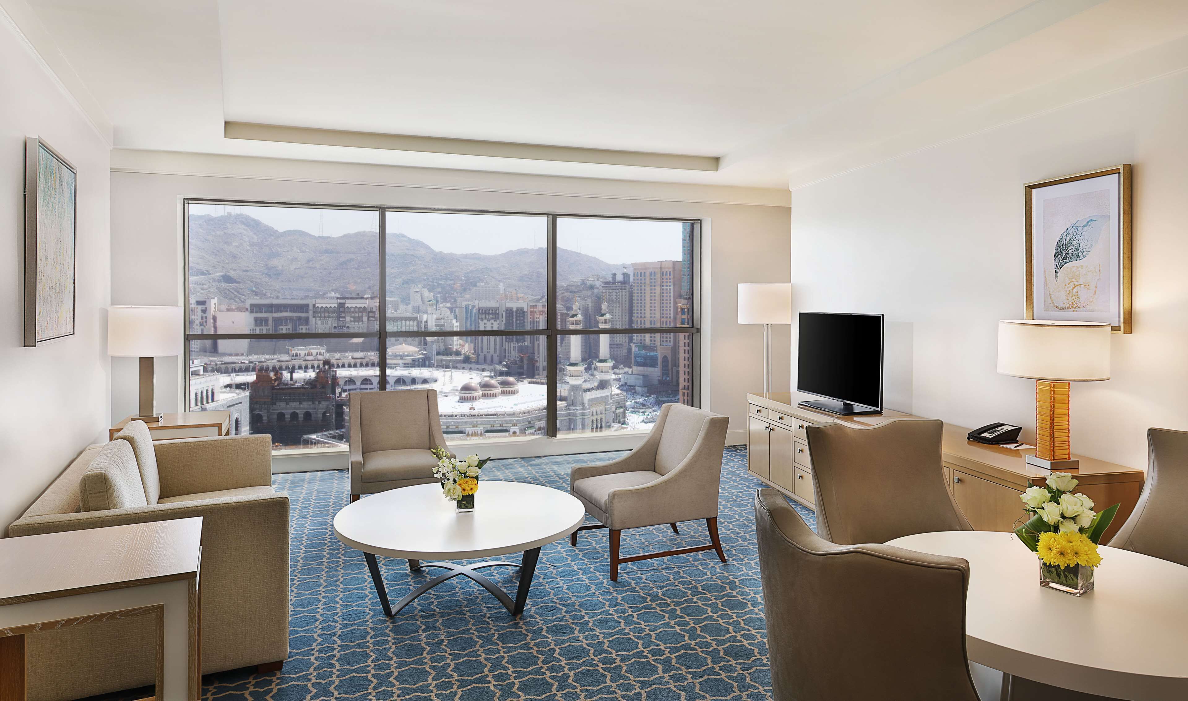 Une chambre d’hôtel dotée d’une grande fenêtre donnant sur un paysage urbain et des montagnes. On y trouve deux canapés, une petite table ronde avec un vase de fleurs et un téléviseur à écran plat posé sur un meuble.