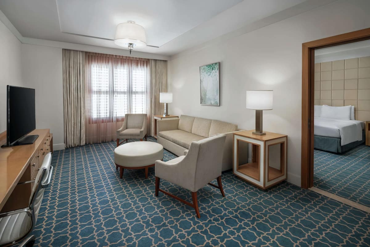A hotel room with a blue patterned carpet, a sofa, two armchairs, a TV, a bedside table with a lamp, and a large mirror.