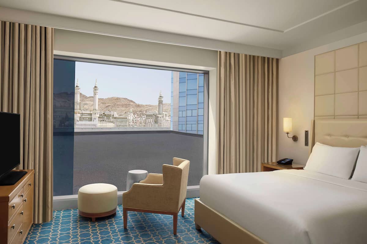 A hotel room with a large bed, a TV, a small table, a chair, and a view of a mosque through a large window.