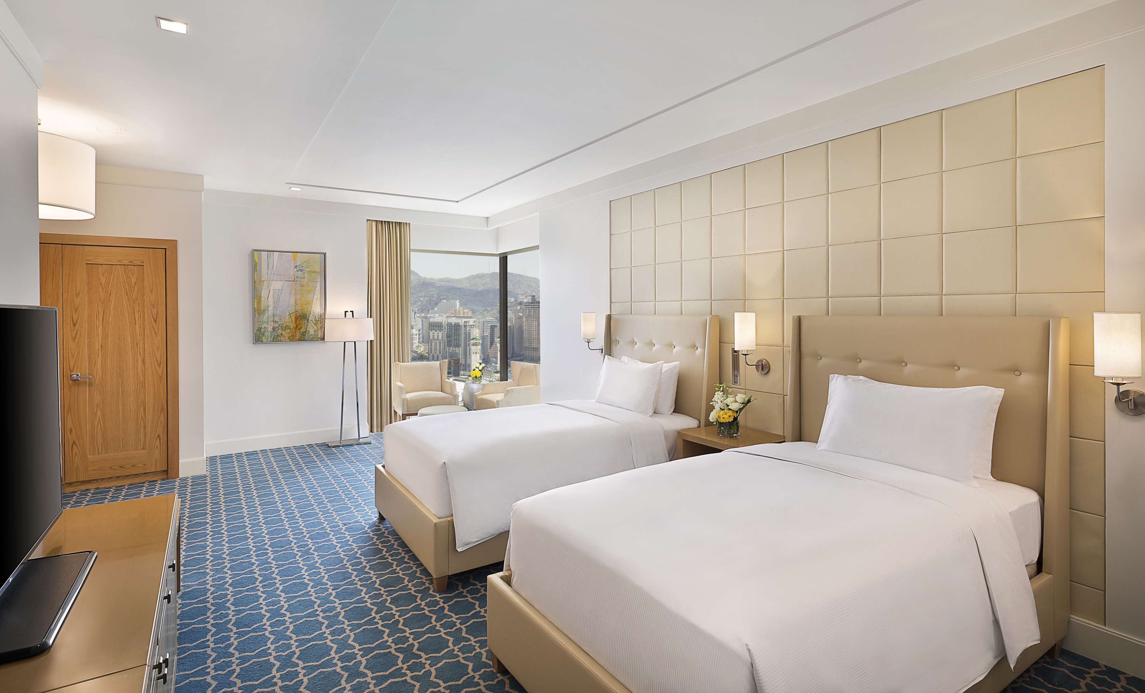 A hotel room with two beds, a TV, a desk, and a view of the city.