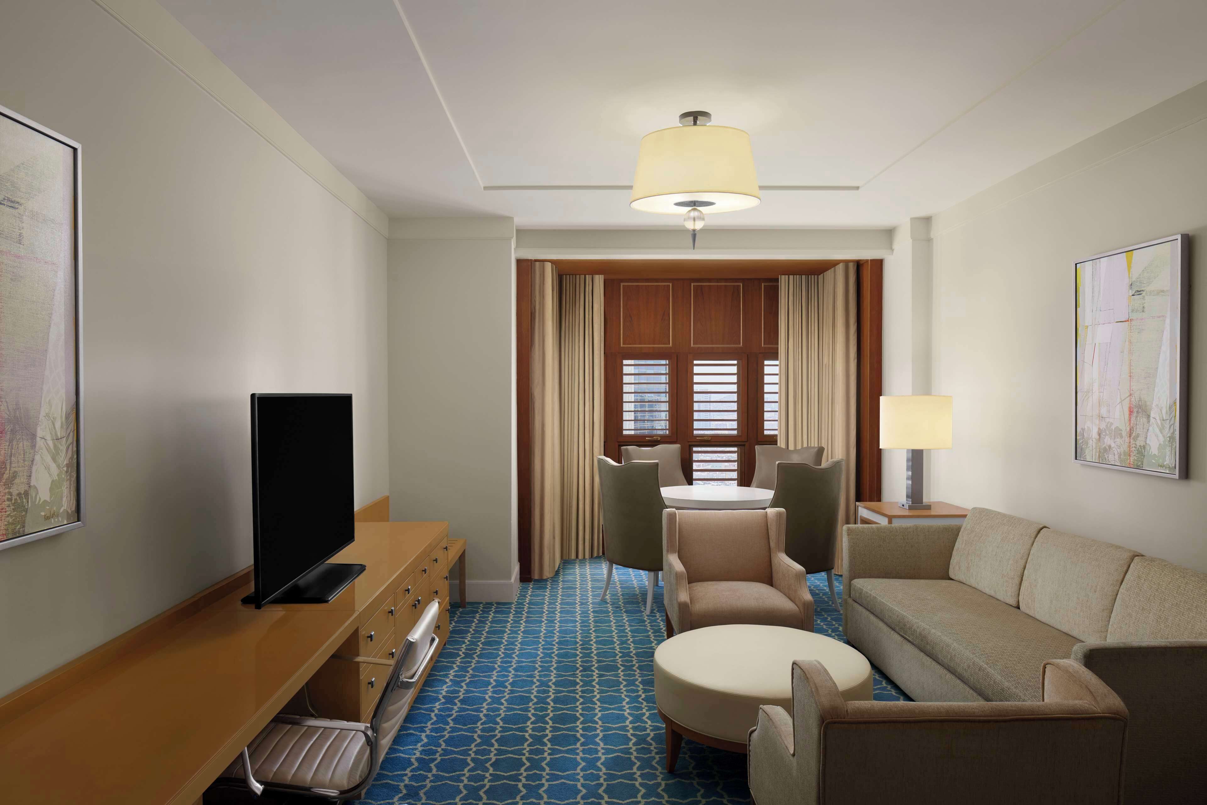 A hotel room with a TV, a desk, a sofa, a chair, a lamp, and a window with curtains.