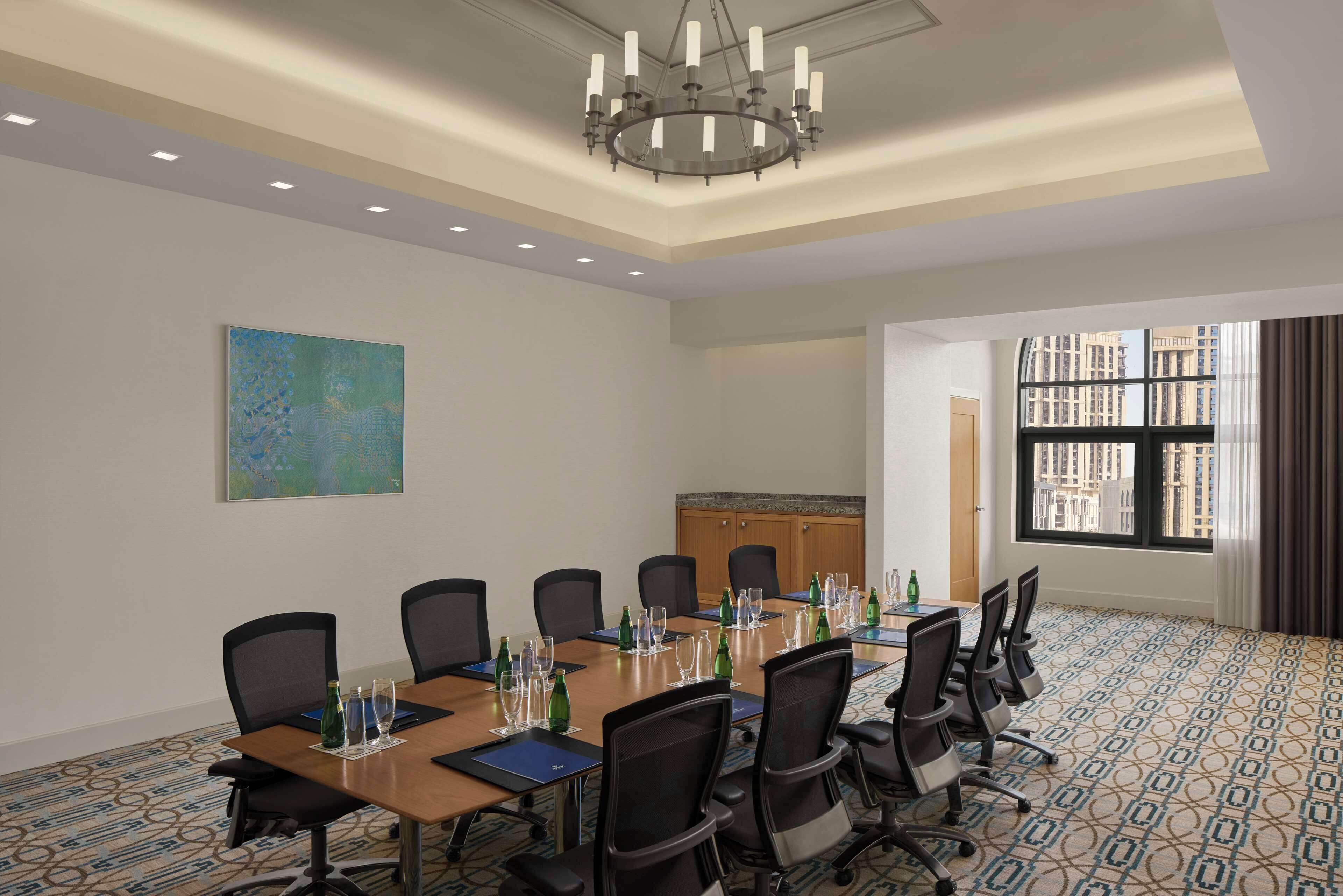 Una sala conferenze con un lungo tavolo, sedie, un lampadario, un quadro e una finestra che si affaccia su edifici.