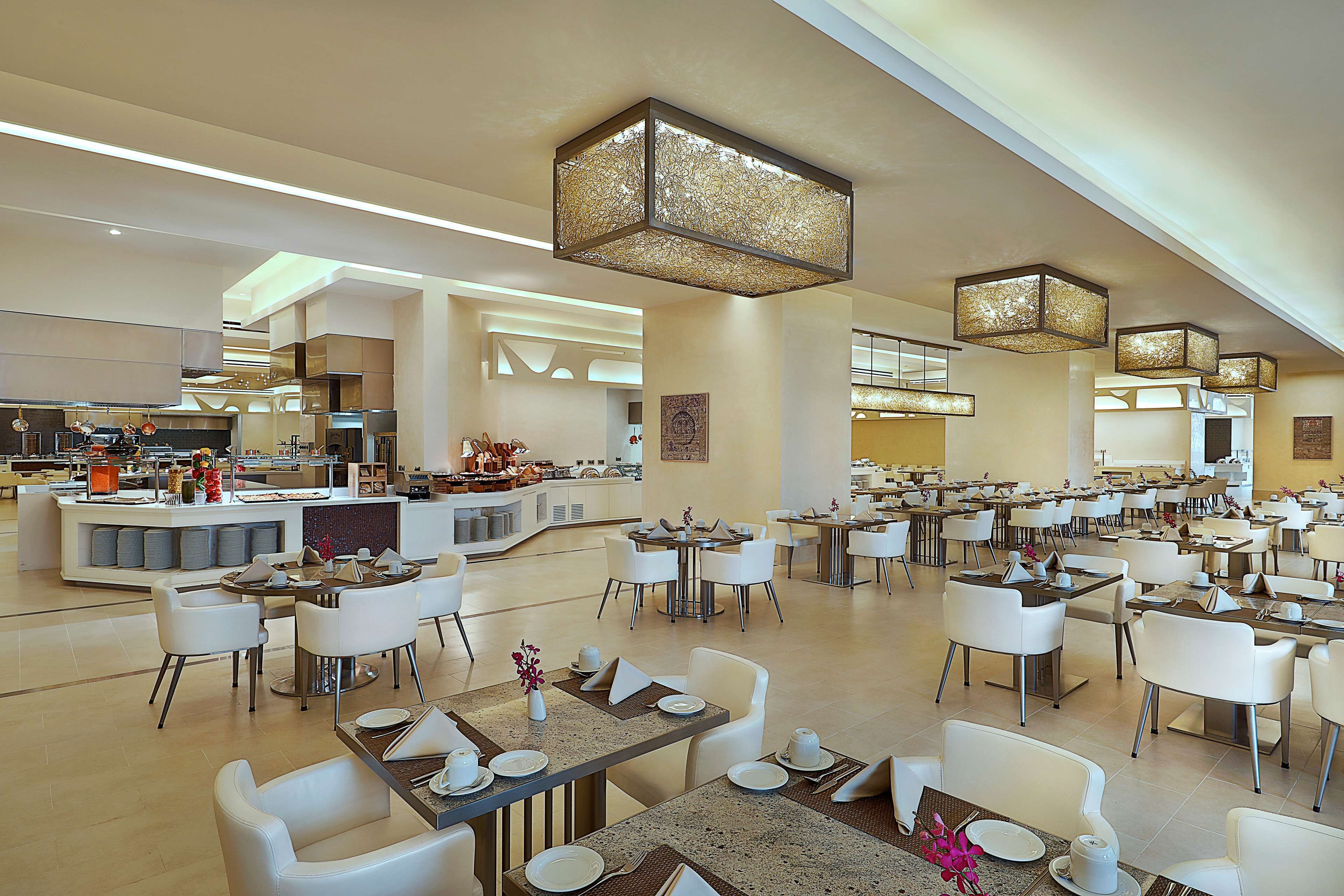 A spacious hotel dining area with multiple tables and chairs, a central bar, and modern lighting fixtures.