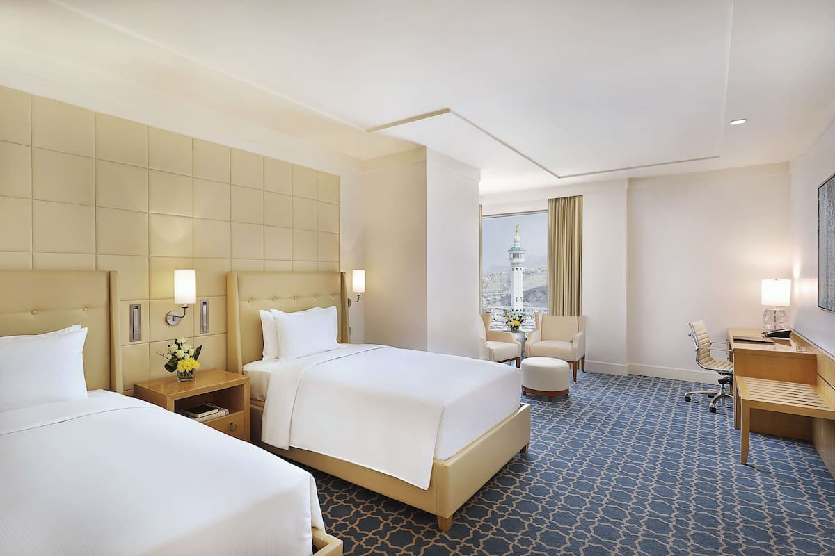 A hotel room with two beds, a desk, a chair, and a view of a tower.