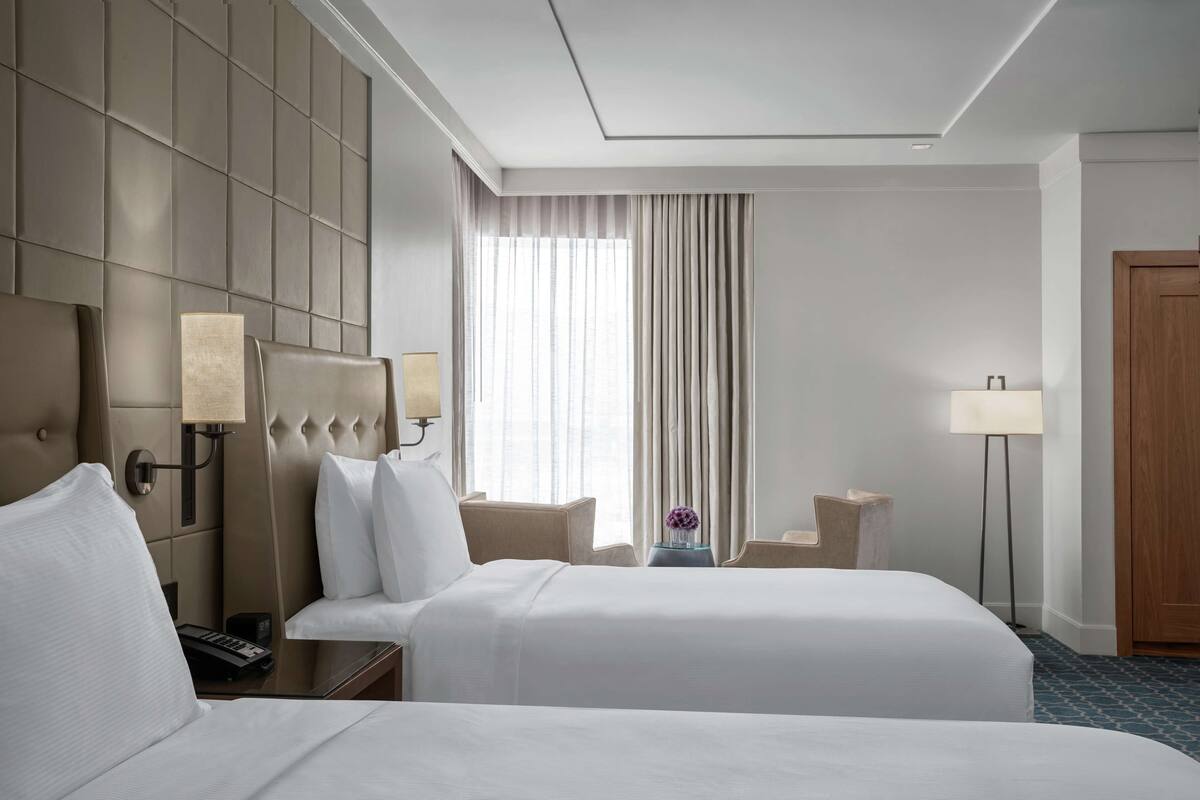 A hotel room with two beds, a large window with curtains, a bedside table with a lamp, and a wooden door.