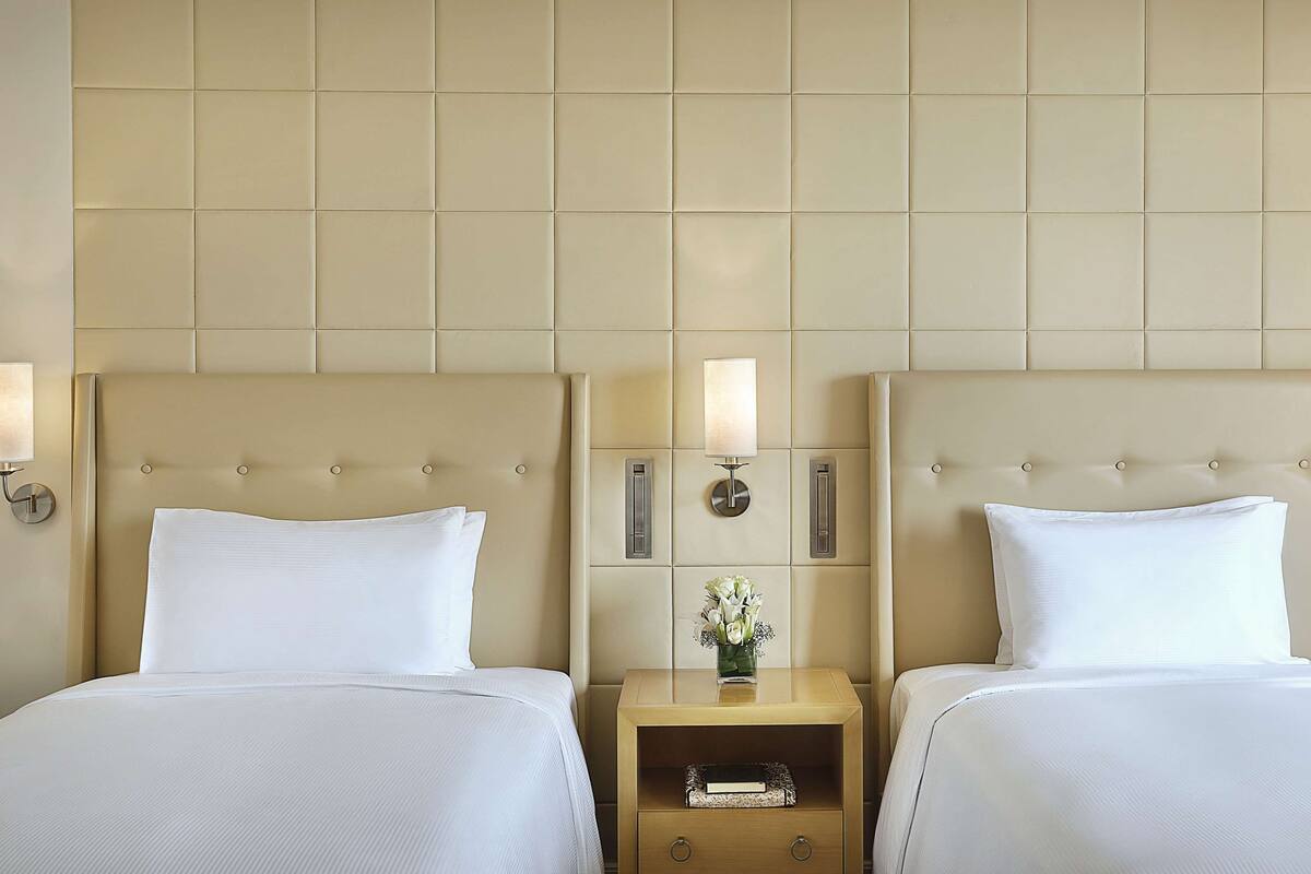 A hotel room with two beds, a nightstand with a lamp, and a wall-mounted light fixture.