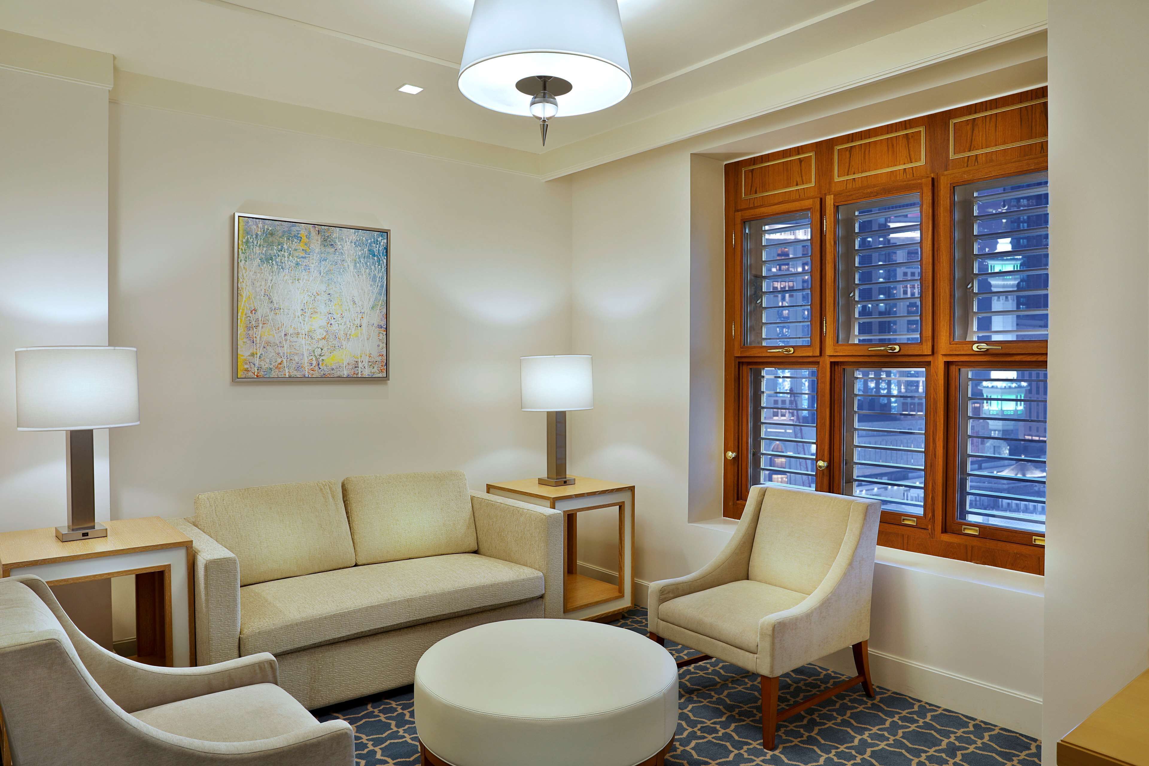 A hotel room with a beige sofa, two armchairs, a round ottoman, a side table with a lamp, a painting on the wall, and a window with blinds.
