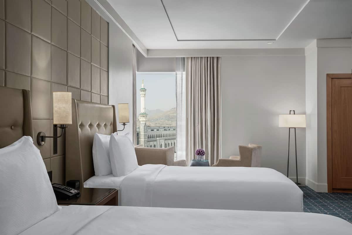 A hotel room with two beds, a large window with a view of a tower, and a view of mountains.