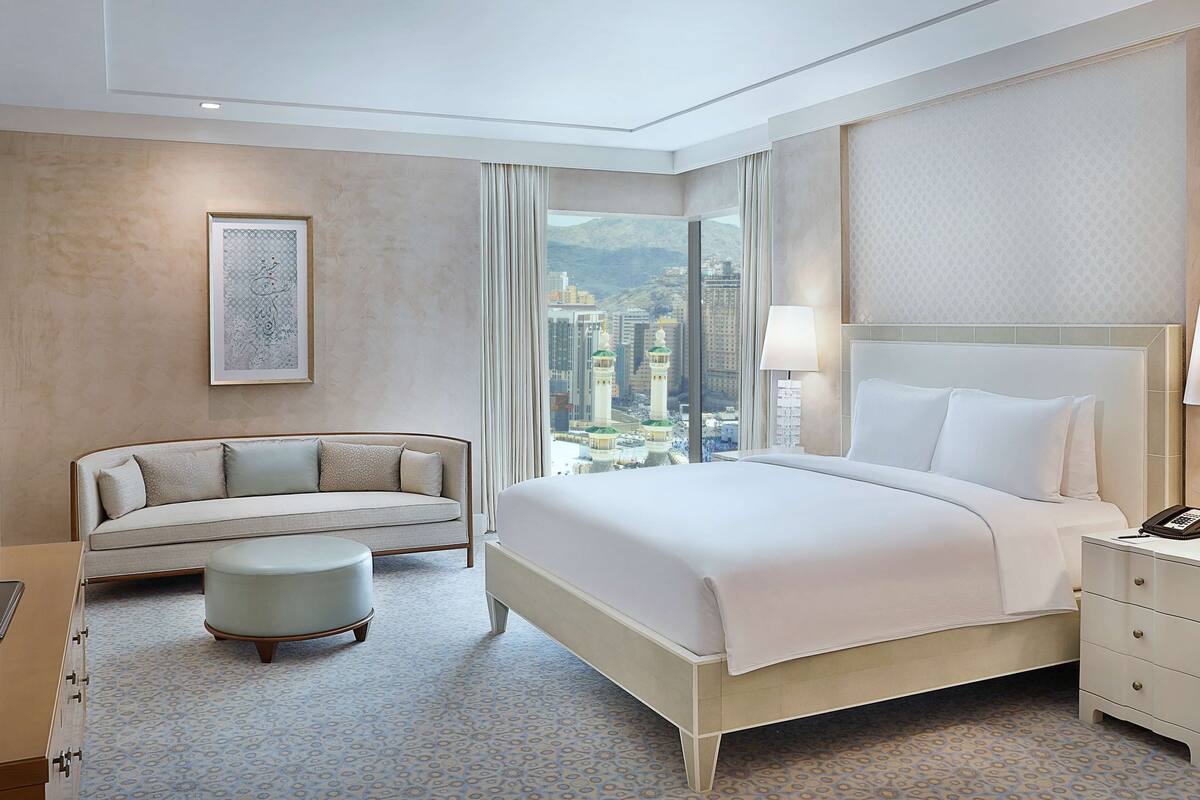 A hotel room with a large bed, a sofa, a small ottoman, a nightstand with a lamp, and a view of a cityscape with tall buildings.